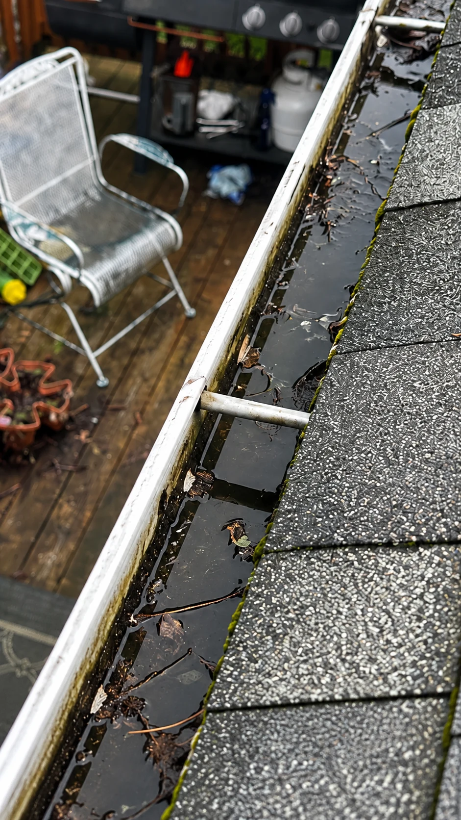 Standing water and debris inside a residential gutter beside asphalt shingles.